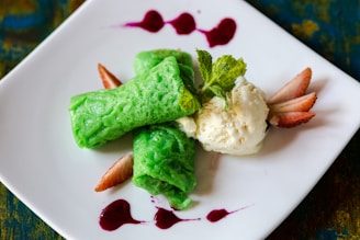 A neatly plated dessert featuring two vibrant green rolled crepes next to a scoop of creamy vanilla ice cream. Fresh mint leaves are placed on top for garnish, while sliced strawberries and drizzles of red sauce add a pop of color on a white square plate.