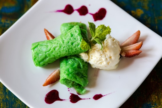 A neatly plated dessert featuring two vibrant green rolled crepes next to a scoop of creamy vanilla ice cream. Fresh mint leaves are placed on top for garnish, while sliced strawberries and drizzles of red sauce add a pop of color on a white square plate.