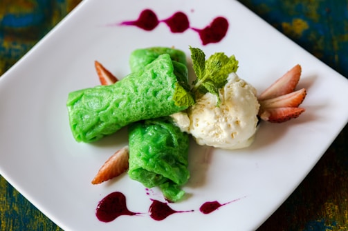 A neatly plated dessert featuring two vibrant green rolled crepes next to a scoop of creamy vanilla ice cream. Fresh mint leaves are placed on top for garnish, while sliced strawberries and drizzles of red sauce add a pop of color on a white square plate.