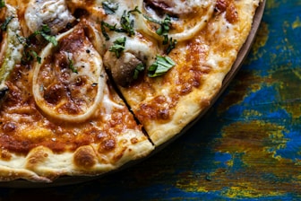 A close-up of a pizza with a crispy, golden-brown crust topped with melted cheese, onion rings, mushrooms, and fresh basil. The surface is bubbly and textured with a rich variety of colors.