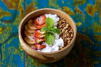 A wooden bowl filled with a colorful assortment of fresh ingredients, including sliced strawberries and bananas, granola, coconut flakes, and vibrant green mint leaves, set against a textured blue and yellow background.