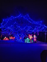 A tree is illuminated with vibrant blue string lights, creating a striking contrast against the dark background. Below the tree, inflatable holiday decorations, including a Santa Claus figure on a motorcycle and a cartoonish reindeer on another vehicle, add a festive touch. Bright, colorful lights enhance these decorations, contributing to a cheerful holiday display.