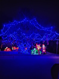 A tree is illuminated with vibrant blue string lights, creating a striking contrast against the dark background. Below the tree, inflatable holiday decorations, including a Santa Claus figure on a motorcycle and a cartoonish reindeer on another vehicle, add a festive touch. Bright, colorful lights enhance these decorations, contributing to a cheerful holiday display.