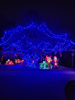 A tree is illuminated with vibrant blue string lights, creating a striking contrast against the dark background. Below the tree, inflatable holiday decorations, including a Santa Claus figure on a motorcycle and a cartoonish reindeer on another vehicle, add a festive touch. Bright, colorful lights enhance these decorations, contributing to a cheerful holiday display.