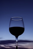 A wine glass filled with red wine is silhouetted against a gradient sky transitioning from blue to light blue with wispy clouds.