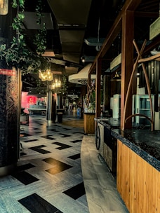 A modern interior space featuring a curved path with geometric patterned flooring, surrounded by wooden structures and lush green plants hanging from the ceiling. Warm lighting from chandeliers creates a cozy and inviting atmosphere.