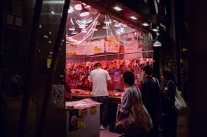 A street-side butcher shop displays various cuts of meat under bright pink lighting. Several people are gathered in front, observing the products offered. Price tags are visible on the meats, and the setting appears to be in an urban area at night, illuminated by overhead lamps.