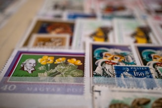 A collection of various postage stamps arranged in a slightly overlapping manner. The stamps feature diverse designs, including portraits of historical figures, botanical illustrations, and colorful artistic depictions. The focal stamp shows a portrait of a man with white hair beside a botanical drawing of yellow flowers on a green background.