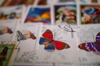 A collection of colorful postage stamps featuring butterfly illustrations. The focus is on a stamp with a vibrant butterfly in red, blue, and brown hues. Other stamps with different butterfly designs and colors are visible in the background, creating an artistic and eclectic display.