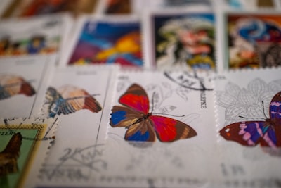 A collection of colorful postage stamps featuring butterfly illustrations. The focus is on a stamp with a vibrant butterfly in red, blue, and brown hues. Other stamps with different butterfly designs and colors are visible in the background, creating an artistic and eclectic display.
