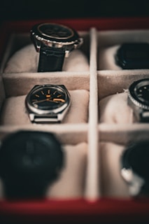 Several watches are neatly arranged in a cushioned watch box with separate compartments. The focus is on a watch with a black strap and another with a silver casing and black face.