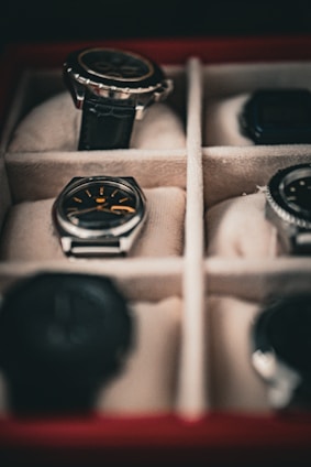 Several watches are neatly arranged in a cushioned watch box with separate compartments. The focus is on a watch with a black strap and another with a silver casing and black face.