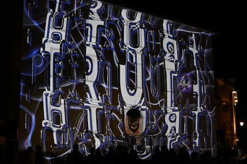 A large wall projection at night features the word 'PERCEPTION' in bold, futuristic, and abstract lettering. The text is intertwined with swirling neon blue and purple lines, creating a dynamic visual effect. Silhouettes of people are seen at the bottom, observing the display.