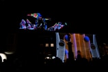 A vibrant projection featuring abstract shapes and patterns illuminates the facade of a building at night. The display includes swirling lines and geometric designs in bright colors, creating a dynamic visual effect against the dark background.