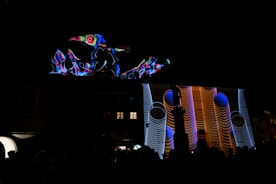 A vibrant projection featuring abstract shapes and patterns illuminates the facade of a building at night. The display includes swirling lines and geometric designs in bright colors, creating a dynamic visual effect against the dark background.