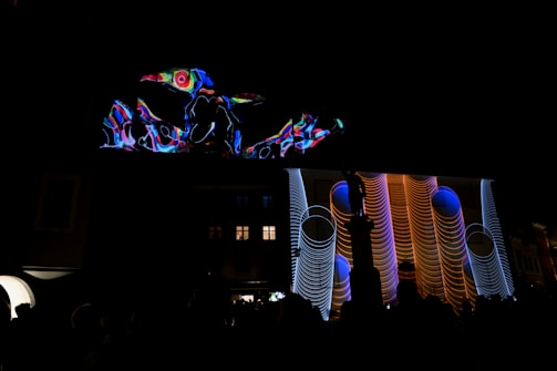 A vibrant projection featuring abstract shapes and patterns illuminates the facade of a building at night. The display includes swirling lines and geometric designs in bright colors, creating a dynamic visual effect against the dark background.