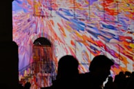 A building facade is illuminated with colorful, abstract light projections. The colors blend in a dynamic burst of reds, blues, yellows, and oranges, creating a vivid and lively atmosphere. Silhouettes of people stand in the foreground, observing the light display.