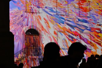 A building facade is illuminated with colorful, abstract light projections. The colors blend in a dynamic burst of reds, blues, yellows, and oranges, creating a vivid and lively atmosphere. Silhouettes of people stand in the foreground, observing the light display.