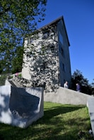A tall, light-gray stucco building with multiple windows sits on a sloped grassy hill under a clear blue sky. The building has a dark, steep roof and casts shadows from nearby trees. Two people are walking up a set of wide stone steps that lead to the building's entrance, alongside a textured concrete wall.