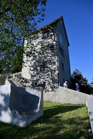 A tall, light-gray stucco building with multiple windows sits on a sloped grassy hill under a clear blue sky. The building has a dark, steep roof and casts shadows from nearby trees. Two people are walking up a set of wide stone steps that lead to the building's entrance, alongside a textured concrete wall.