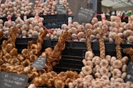 A market stall displays bundles of garlic and onions organized neatly in black crates. Some of the garlic is braided and labeled with signs indicating varieties and prices. The overall setup suggests a focus on quality produce, possibly from France.