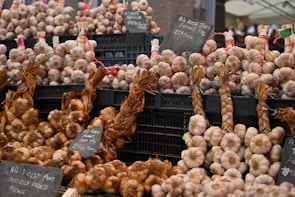 A market stall displays bundles of garlic and onions organized neatly in black crates. Some of the garlic is braided and labeled with signs indicating varieties and prices. The overall setup suggests a focus on quality produce, possibly from France.