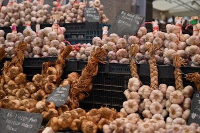 A market stall displays bundles of garlic and onions organized neatly in black crates. Some of the garlic is braided and labeled with signs indicating varieties and prices. The overall setup suggests a focus on quality produce, possibly from France.