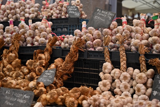 A market stall displays bundles of garlic and onions organized neatly in black crates. Some of the garlic is braided and labeled with signs indicating varieties and prices. The overall setup suggests a focus on quality produce, possibly from France.