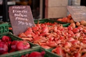 A market stall displays a variety of vegetables, including a large pile of pink shallots in rectangular green crates. A chalkboard sign details the product and its price in French, indicating its origin from Brittany, France. In the background, there are earthy brown potatoes visible in another crate.