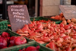 A market stall displays a variety of vegetables, including a large pile of pink shallots in rectangular green crates. A chalkboard sign details the product and its price in French, indicating its origin from Brittany, France. In the background, there are earthy brown potatoes visible in another crate.