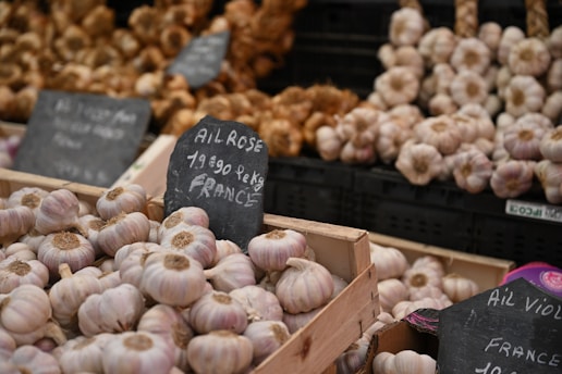 A market stall displays multiple baskets filled with garlic bulbs. Chalkboard signs are present, indicating details about the garlic, including price and origin. The setting suggests an organized and abundant display of produce, with various types of garlic visible in the background.