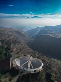 The image captures a large satellite dish or radio telescope situated in a mountainous area with barren, brown earth and sparse vegetation. To the right of the telescope, there is a winding path leading up the mountains. In the background, a misty landscape with a distant mountain range is visible, partly covered by clouds. The sky is clear and blue, providing a contrast to the earthy tones of the landscape. There is a building adjacent to the left side of the telescope.