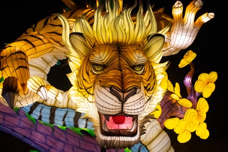 An intricate, illuminated sculpture featuring a stylized tiger with expressive eyes and an open mouth, surrounded by vibrant yellow flower shapes. The tiger appears to be part of a larger, winding form, with detailed patterns and luminous colors.