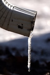 A close-up view of a metal downspout with a long translucent icicle hanging from its tip, casting a shadow against a blurred, cold background.