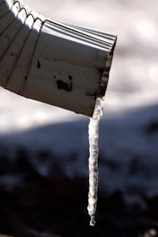 A close-up view of a metal downspout with a long translucent icicle hanging from its tip, casting a shadow against a blurred, cold background.