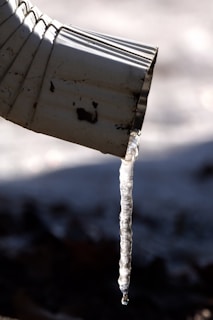 A close-up view of a metal downspout with a long translucent icicle hanging from its tip, casting a shadow against a blurred, cold background.