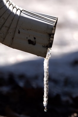 A close-up view of a metal downspout with a long translucent icicle hanging from its tip, casting a shadow against a blurred, cold background.