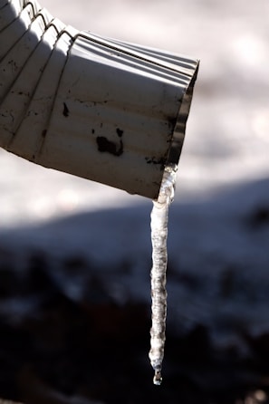 A close-up view of a metal downspout with a long translucent icicle hanging from its tip, casting a shadow against a blurred, cold background.