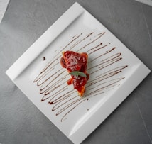 A slice of cake topped with red berry sauce is placed on a large white square plate. The plate is artistically decorated with drizzles of chocolate sauce. The background is a textured grey surface.