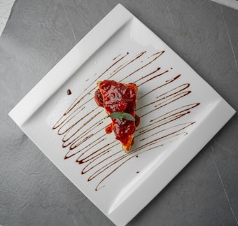 A slice of cake topped with red berry sauce is placed on a large white square plate. The plate is artistically decorated with drizzles of chocolate sauce. The background is a textured grey surface.