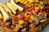 A vibrant and appetizing spread of grilled seafood, including shrimp, surrounded by colorful roasted vegetables such as bell peppers, onions, and eggplant on a wooden board. Accompanying the main dish are crispy, golden plantain slices and thick-cut yucca fries.