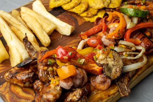 A vibrant and appetizing spread of grilled seafood, including shrimp, surrounded by colorful roasted vegetables such as bell peppers, onions, and eggplant on a wooden board. Accompanying the main dish are crispy, golden plantain slices and thick-cut yucca fries.