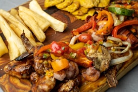 A vibrant and appetizing spread of grilled seafood, including shrimp, surrounded by colorful roasted vegetables such as bell peppers, onions, and eggplant on a wooden board. Accompanying the main dish are crispy, golden plantain slices and thick-cut yucca fries.