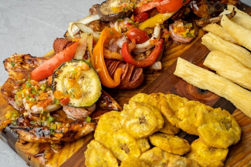 A wooden platter features a variety of grilled vegetables and meats, including zucchini, red bell peppers, and onions, topped with a colorful salsa. Accompanying the main dish are fried plantains and crispy yuca fries.