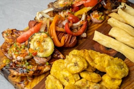 A wooden platter features a variety of grilled vegetables and meats, including zucchini, red bell peppers, and onions, topped with a colorful salsa. Accompanying the main dish are fried plantains and crispy yuca fries.