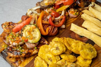 A wooden platter features a variety of grilled vegetables and meats, including zucchini, red bell peppers, and onions, topped with a colorful salsa. Accompanying the main dish are fried plantains and crispy yuca fries.