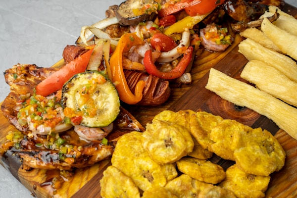 A wooden platter features a variety of grilled vegetables and meats, including zucchini, red bell peppers, and onions, topped with a colorful salsa. Accompanying the main dish are fried plantains and crispy yuca fries.