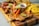 A wooden board featuring a variety of grilled and fried foods including grilled chicken topped with colorful bell peppers and lime, fried plantains, sausages, and a small bowl of dipping sauce.