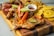 A wooden board featuring a variety of grilled and fried foods including grilled chicken topped with colorful bell peppers and lime, fried plantains, sausages, and a small bowl of dipping sauce.