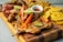 A wooden board featuring a variety of grilled and fried foods including grilled chicken topped with colorful bell peppers and lime, fried plantains, sausages, and a small bowl of dipping sauce.
